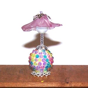 May include: A miniature lamp with a pink flower-shaped shade and a colorful, jeweled base. The lamp has a silver stem and base, and the base is adorned with iridescent, colorful gems. The lamp is set on a wooden surface.
