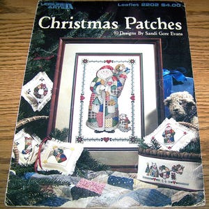 May include: A Leisure Arts craft leaflet titled Christmas Patches, 10 Designs By Sandi Gore Evans, featuring a cross-stitch pattern of a Santa Claus figure in a patchwork coat, surrounded by smaller embroidered ornaments and holiday motifs.