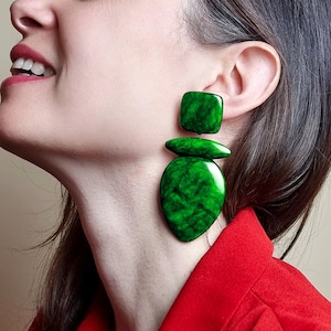 May include: A large statement earring featuring three distinct, glossy, emerald green components with a marbled texture. The design consists of a square top piece, a horizontal oval middle section, and a large, teardrop-shaped bottom element.