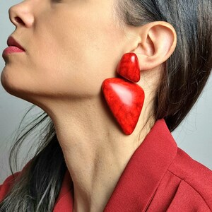 May include: A pair of large, statement earrings featuring two distinct, smooth, red pieces with a marbled texture. The top piece is a smaller, rounded triangle shape, while the bottom piece is a larger, elongated, asymmetrical triangular shape.