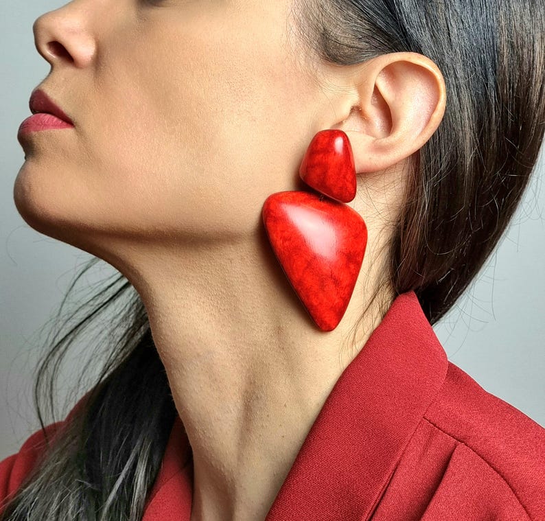 May include: A pair of large, statement earrings featuring two distinct, smooth, red pieces with a marbled texture. The top piece is a smaller, rounded triangle shape, while the bottom piece is a larger, elongated, asymmetrical triangular shape.
