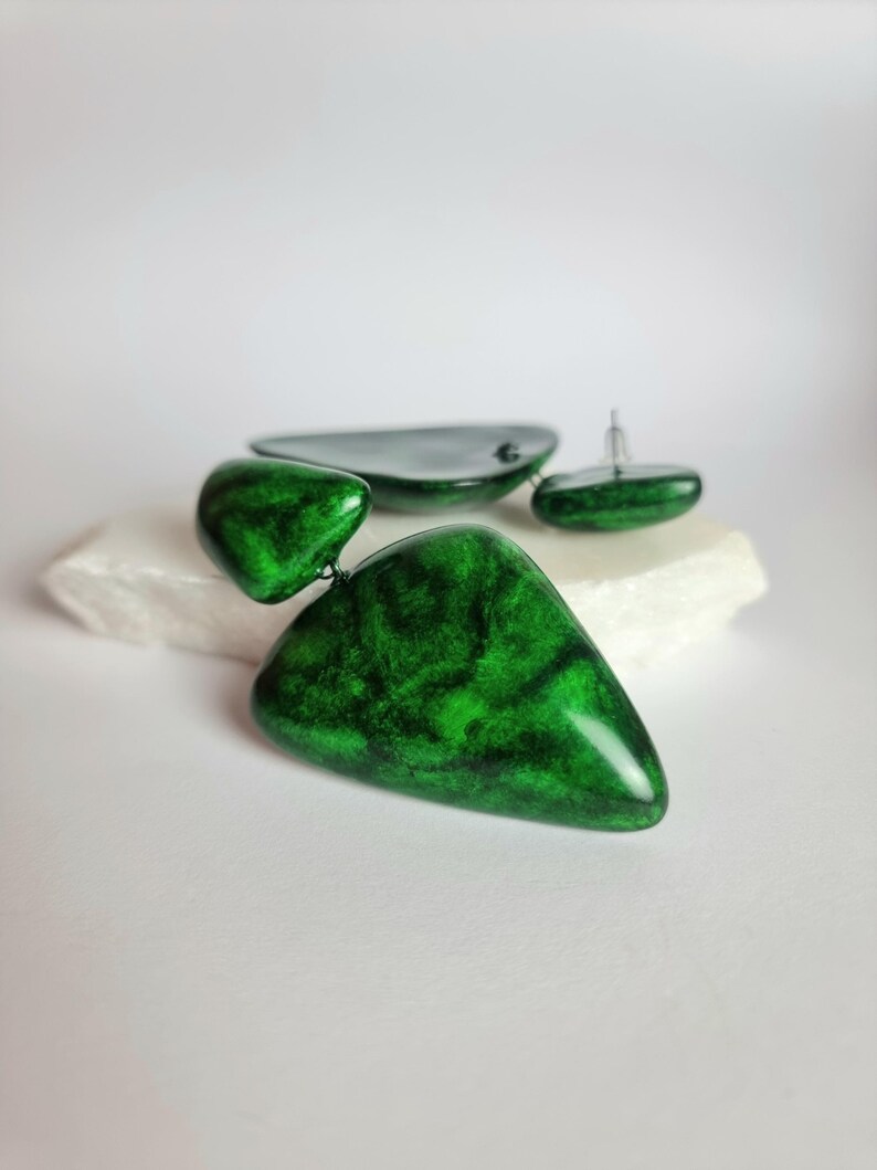 May include: A pair of drop earrings featuring two connected, irregular triangular shapes in a glossy, marbled emerald green finish. The earrings have a smooth, polished surface with dark green veining throughout and include silver-toned earring posts.