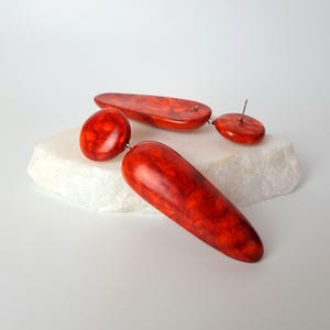 Op de afbeelding: Een paar hangende oorbellen met een levendig oranje en rood gemarmerd patroon. Elke oorbel bestaat uit een kleine ronde knop verbonden met een grotere, langwerpige, gladde, druppelvormige hanger. De afwerking is glanzend en gepolijst.