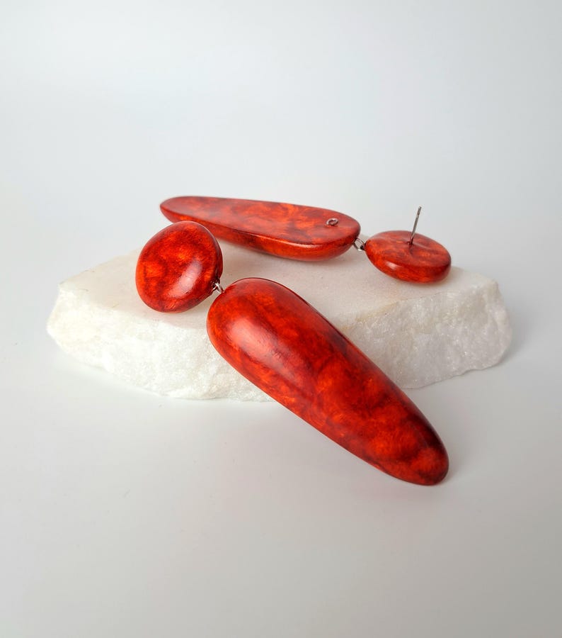 Op de afbeelding: Een paar hangende oorbellen met een levendig oranje en rood gemarmerd patroon. Elke oorbel bestaat uit een kleine ronde knop verbonden met een grotere, langwerpige, gladde, druppelvormige hanger. De afwerking is glanzend en gepolijst.