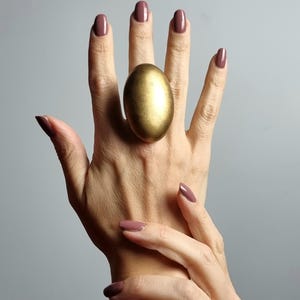 K&ouml;nnte beinhalten: Ein gro&szlig;er, goldfarbener, eif&ouml;rmiger Ring wird an einer Hand mit passendem mauvefarbenem Nagellack getragen. Der Ring ist der Blickfang, mit einer strukturierten Oberfl&auml;che und einer markanten, abgerundeten Form. Der Hintergrund ist einfarbiges, neutrales Grau.