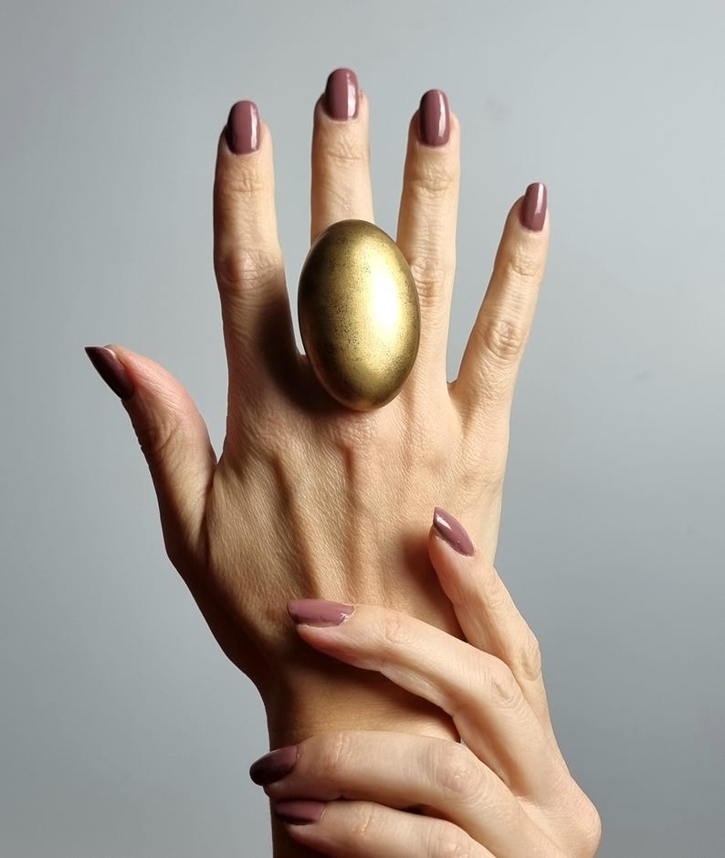 K&ouml;nnte beinhalten: Ein gro&szlig;er, goldfarbener, eif&ouml;rmiger Ring wird an einer Hand mit passendem mauvefarbenem Nagellack getragen. Der Ring ist der Blickfang, mit einer strukturierten Oberfl&auml;che und einer markanten, abgerundeten Form. Der Hintergrund ist einfarbiges, neutrales Grau.