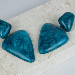 May include: A pair of drop earrings featuring two teal-colored, marbled, triangular-shaped components connected by a small metal link. The surface of each piece displays a smooth, polished finish with deep blue and green swirling patterns.