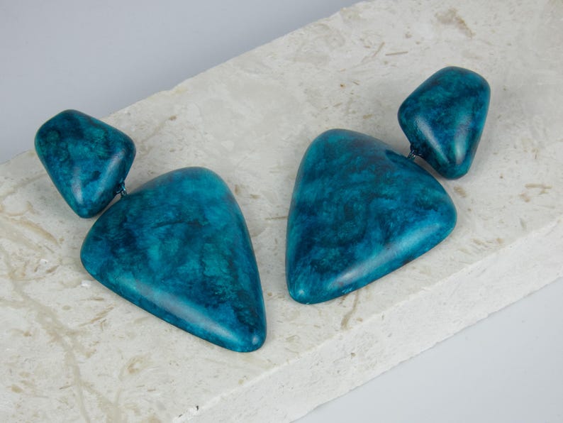 May include: A pair of drop earrings featuring two teal-colored, marbled, triangular-shaped components connected by a small metal link. The surface of each piece displays a smooth, polished finish with deep blue and green swirling patterns.