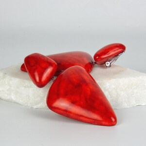 May include: A pair of clip-on earrings featuring three smooth, polished, bright red triangular components of varying sizes connected in a cascading drop design. The vibrant red material has a marbled texture with subtle darker red veining throughout.