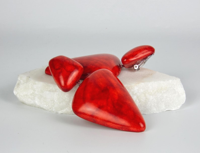 May include: A pair of clip-on earrings featuring three smooth, polished, bright red triangular components of varying sizes connected in a cascading drop design. The vibrant red material has a marbled texture with subtle darker red veining throughout.
