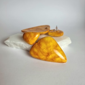 May include: A pair of drop earrings featuring smooth, triangular-shaped components in a vibrant mustard yellow with marbled amber-toned swirls. The earrings have a glossy finish and are constructed with a post-back closure for secure attachment.
