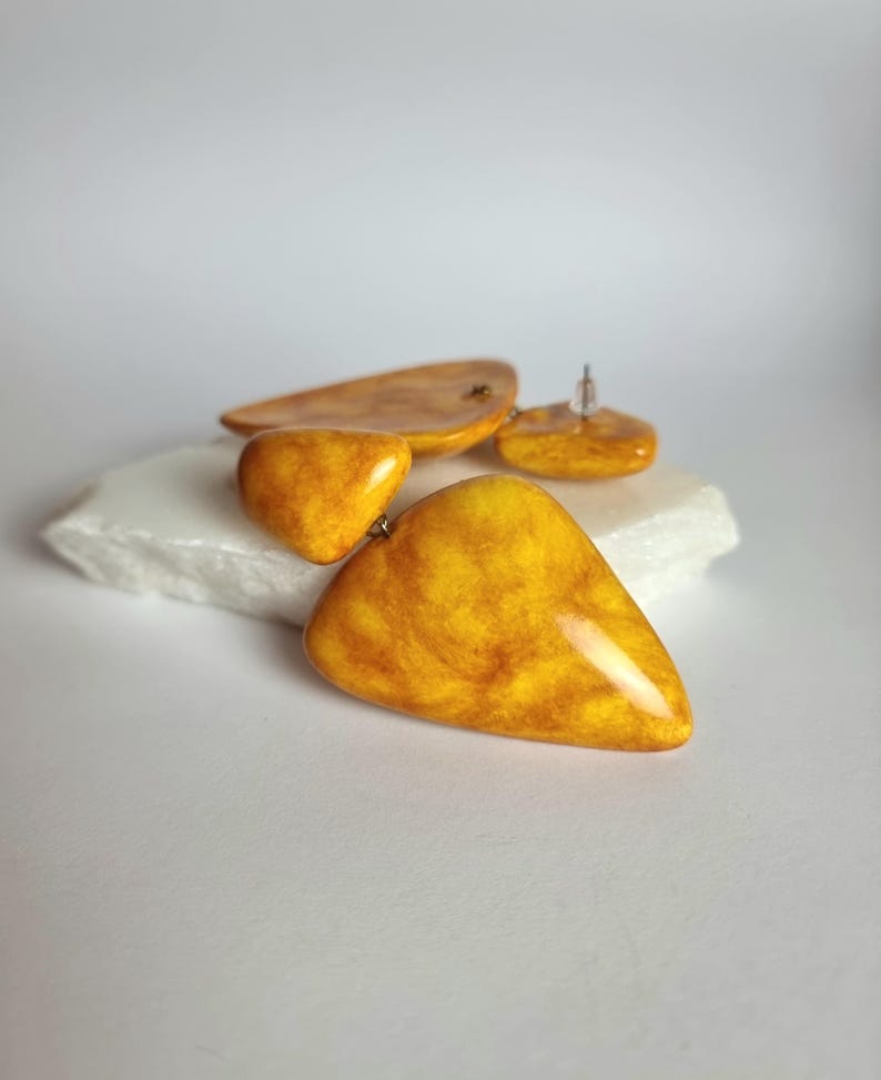May include: A pair of drop earrings featuring smooth, triangular-shaped components in a vibrant mustard yellow with marbled amber-toned swirls. The earrings have a glossy finish and are constructed with a post-back closure for secure attachment.