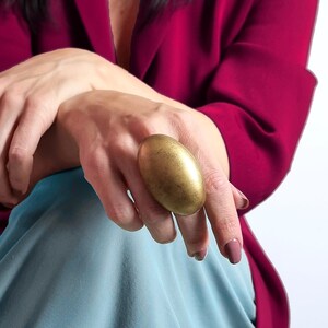 K&ouml;nnte beinhalten: Ein gro&szlig;er, ovaler Goldring. Der Ring wird an einem Finger getragen, vor dem Hintergrund eines magentafarbenen Blazers und hellblauem Stoff. Die Oberfl&auml;che des Rings hat ein strukturiertes, gealtertes Aussehen.