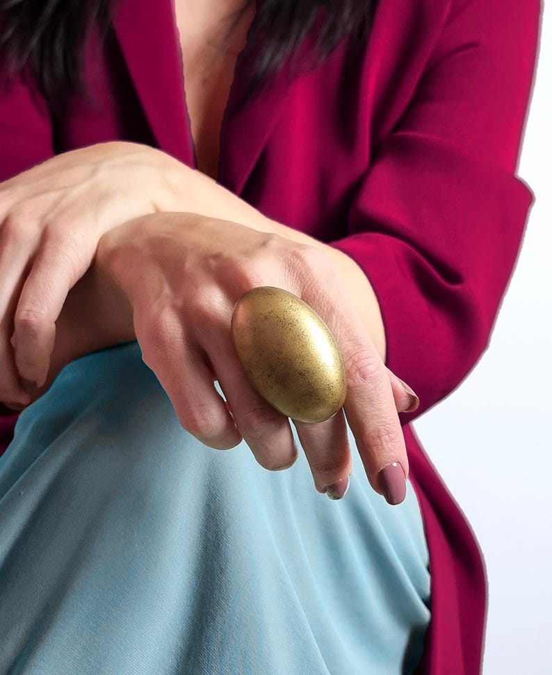 K&ouml;nnte beinhalten: Ein gro&szlig;er, ovaler Goldring. Der Ring wird an einem Finger getragen, vor dem Hintergrund eines magentafarbenen Blazers und hellblauem Stoff. Die Oberfl&auml;che des Rings hat ein strukturiertes, gealtertes Aussehen.