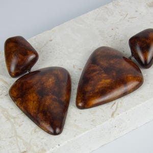May include: A pair of drop earrings featuring two smooth, polished brown pieces with a mottled, tortoiseshell-like pattern. Each earring consists of a smaller rounded top piece connected to a larger, triangular bottom piece with rounded edges.