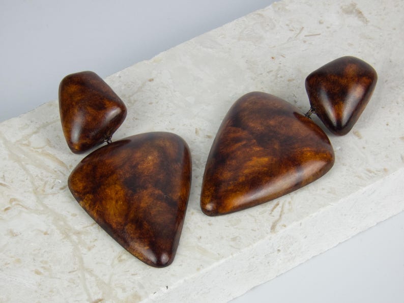 May include: A pair of drop earrings featuring two smooth, polished brown pieces with a mottled, tortoiseshell-like pattern. Each earring consists of a smaller rounded top piece connected to a larger, triangular bottom piece with rounded edges.