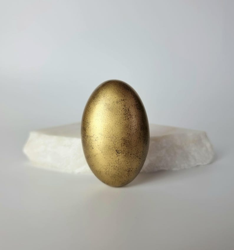 K&ouml;nnte beinhalten: Ein goldfarbenes, eif&ouml;rmiges Objekt mit strukturierter Oberfl&auml;che. Das Ei steht auf einem wei&szlig;en, unregelm&auml;&szlig;ig geformten Stein vor einem wei&szlig;en Hintergrund. Das Ei hat eine metallische Oberfl&auml;che mit dunkleren braunen Akzenten.