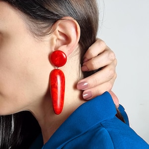 Op de afbeelding: Een paar statement oorbellen met een gladde, glanzende rode ronde knop verbonden aan een lange, taps toelopende, druppelvormige hanger in een bijpassende levendige rode tint met een subtiele gemarmerde textuur over het gepolijste oppervlak.