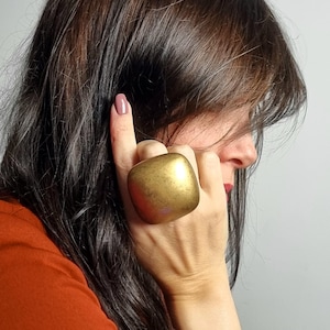 May include: A large, square-shaped ring with a smooth, matte gold-toned finish. The ring features rounded corners and a substantial, domed surface that covers a significant portion of the finger, showcasing a metallic texture and warm golden hue.