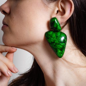 May include: A pair of statement earrings featuring two distinct, polished green resin components. The top piece is a small, rounded organic shape, while the bottom piece is a larger, elongated triangular form with a swirling, marbled texture.