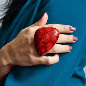 Op de afbeelding: Een grote, gladde, druppelvormige ring met een levendige, gemarmerde rode afwerking met donkerdere karmozijnrode ondertonen. Het opvallende sieraad heeft een gepolijst, glanzend oppervlak en een afgeronde, organische vorm.