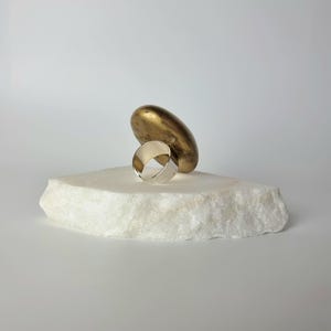 K&ouml;nnte beinhalten: Ein silberner Ring mit breitem Band liegt auf einem wei&szlig;en, strukturierten Stein. Eine goldfarbene, l&auml;ngliche Skulptur ruht hinter dem Ring. Der Ring ist ein schlichtes, modernes Design, das Licht reflektiert. Die Komposition ist minimalistisch und elegant.