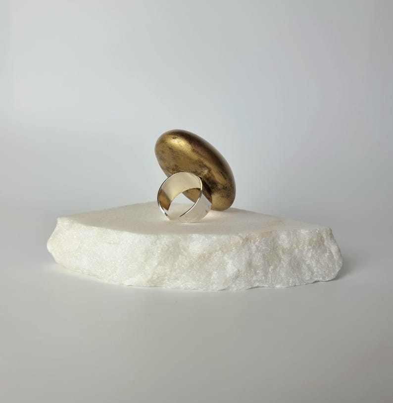 K&ouml;nnte beinhalten: Ein silberner Ring mit breitem Band liegt auf einem wei&szlig;en, strukturierten Stein. Eine goldfarbene, l&auml;ngliche Skulptur ruht hinter dem Ring. Der Ring ist ein schlichtes, modernes Design, das Licht reflektiert. Die Komposition ist minimalistisch und elegant.