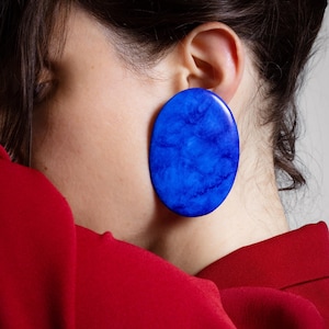 Puede incluir: Primer plano de un pendiente azul grande, de forma ovalada. El pendiente tiene una textura veteada y se encuentra sobre una prenda roja y un fondo neutro. El pendiente es una pieza llamativa.