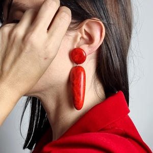 Op de afbeelding: Een paar hangende oorbellen met een gladde, glanzende, felrode afwerking. Elke oorbel bestaat uit een kleine ronde knop aan de bovenkant, verbonden met een grotere, langwerpige, druppelvormige hanger die verticaal aan de oorlel hangt.