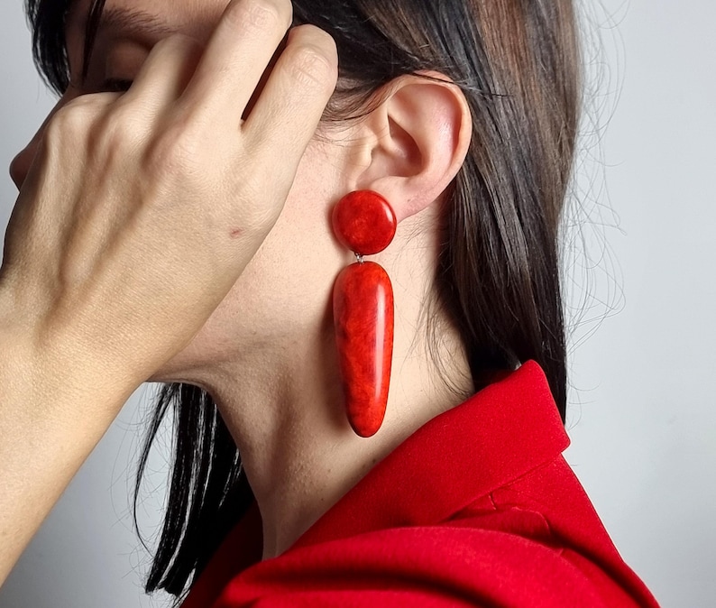 Op de afbeelding: Een paar hangende oorbellen met een gladde, glanzende, felrode afwerking. Elke oorbel bestaat uit een kleine ronde knop aan de bovenkant, verbonden met een grotere, langwerpige, druppelvormige hanger die verticaal aan de oorlel hangt.