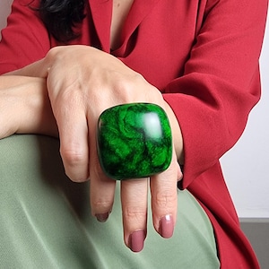 May include: A large, square-shaped ring featuring a polished, marbled surface with deep emerald green and black swirls. The ring has a smooth, rounded edge design and a glossy finish that reflects light, creating a bold, prominent appearance on the finger.