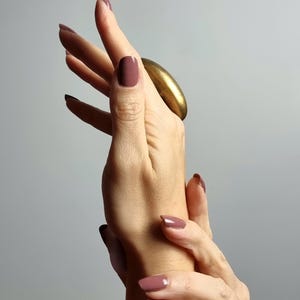 K&ouml;nnte beinhalten: Eine Hand mit mauvefarbenem Nagellack und einem gro&szlig;en, goldfarbenen Ring. Die N&auml;gel sind mit einer einfarbigen Farbe lackiert. Der Ring ist oval und hat eine glatte, metallische Oberfl&auml;che. Der Hintergrund ist ein weiches, neutrales Grau.