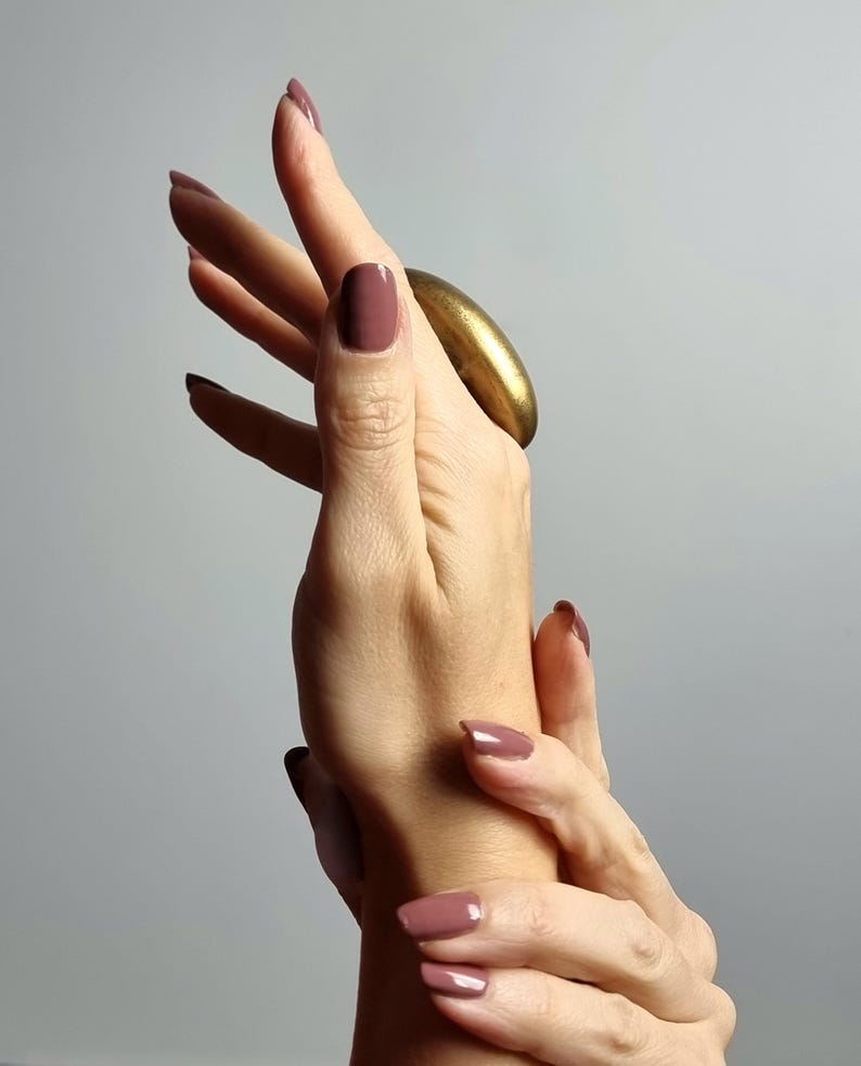 K&ouml;nnte beinhalten: Eine Hand mit mauvefarbenem Nagellack und einem gro&szlig;en, goldfarbenen Ring. Die N&auml;gel sind mit einer einfarbigen Farbe lackiert. Der Ring ist oval und hat eine glatte, metallische Oberfl&auml;che. Der Hintergrund ist ein weiches, neutrales Grau.