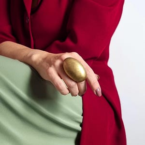 K&ouml;nnte beinhalten: Ein gro&szlig;er, ovaler goldener Ring. Der Ring wird an einer Hand getragen, mit einer roten Jacke und einem gr&uuml;nen Oberteil im Hintergrund. Die Oberfl&auml;che des Rings hat ein strukturiertes Aussehen und f&auml;ngt das Licht ein.