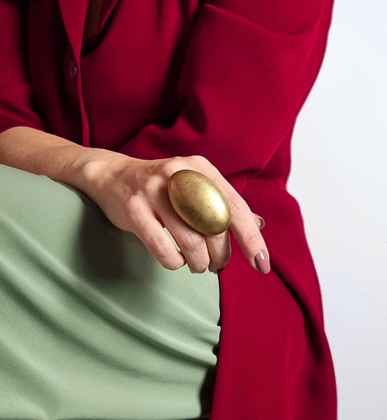 K&ouml;nnte beinhalten: Ein gro&szlig;er, ovaler goldener Ring. Der Ring wird an einer Hand getragen, mit einer roten Jacke und einem gr&uuml;nen Oberteil im Hintergrund. Die Oberfl&auml;che des Rings hat ein strukturiertes Aussehen und f&auml;ngt das Licht ein.