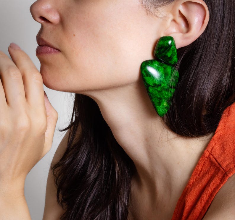 May include: A large, statement earring featuring two distinct, polished green pieces with a marbled texture. The top section is a smaller, rounded shape, while the bottom section is a larger, elongated, triangular form with smooth, glossy surfaces.