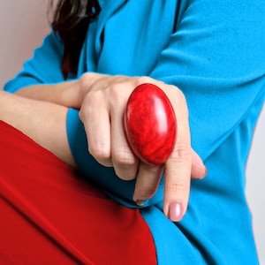 Puede incluir: Un anillo grande de forma ovalada con una piedra roja lisa y pulida con vetas jaspeadas de color rojo oscuro. El anillo se lleva en un dedo, sobre una prenda superior azul brillante y una prenda roja, con un diseño redondeado y brillante.