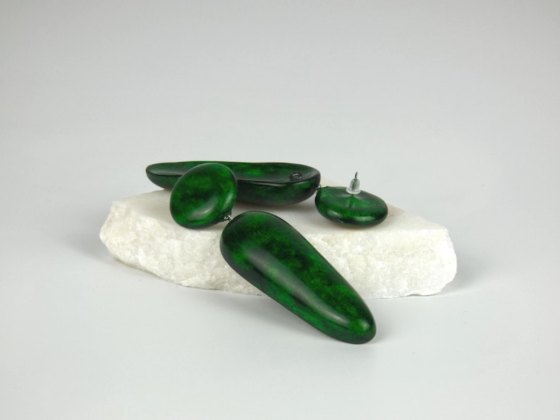 Op de afbeelding: Een paar donkergroene oorbellen met een gladde, gepolijste afwerking en een gevlekte textuur. Elke oorbel bestaat uit een ronde knop verbonden met een lange, druppelvormige hanger. De oorbellen hebben een glanzend uiterlijk en zilverkleurige stekers.