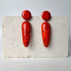 Op de afbeelding: Een paar oorbellen met een ronde knop en een lange, taps toelopende druppelvormige hanger. De oorbellen hebben een gepolijste, glanzende afwerking met een gemarmerd patroon in dieprode en oranje tinten over het gladde oppervlak.