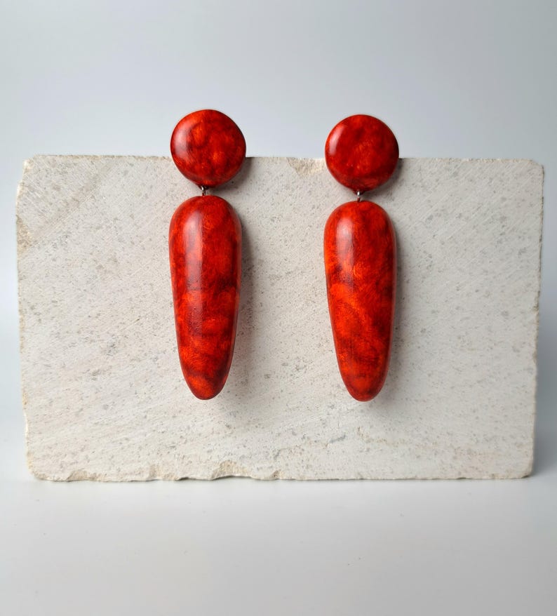 Op de afbeelding: Een paar oorbellen met een ronde knop en een lange, taps toelopende druppelvormige hanger. De oorbellen hebben een gepolijste, glanzende afwerking met een gemarmerd patroon in dieprode en oranje tinten over het gladde oppervlak.