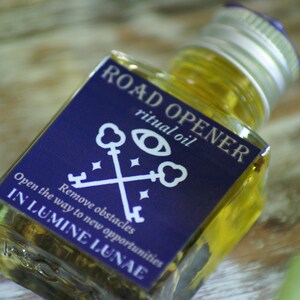 May include: A square glass bottle filled with yellow liquid, labelled "ROAD OPENER ritual oil." The dark blue label features white text and key graphics. The text on the label reads "Open the way to new opportunities" and "IN LUMINE LUNAE."