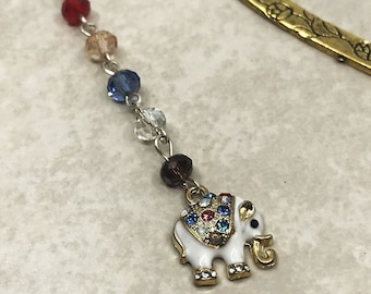 May include: A gold bookmark with a floral design and a white elephant charm with colourful rhinestones. The elephant charm is attached to a chain with colourful beads.