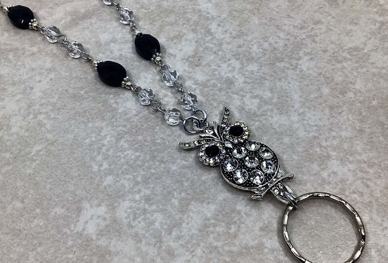 May include: A silver owl charm with clear and black rhinestones, attached to a black and clear beaded lanyard with a silver ring.