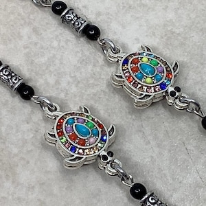 May include: Silver-toned turtle charm bracelets with colorful gemstone accents. Each turtle has a turquoise center stone and is surrounded by a rainbow of gems. The bracelets also feature black beads and decorative silver links.