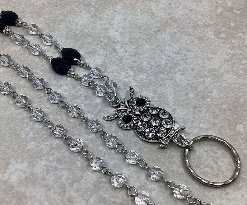 May include: A silver owl charm with black rhinestones hangs from a clear crystal beaded lanyard with a silver ring.