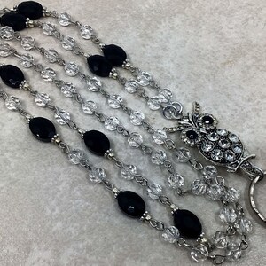 May include: Multi-strand beaded lanyard with black and clear beads.  The lanyard features an ornate owl-shaped badge holder with clear rhinestone accents. The metal chain and clasp are silver-toned.  The black beads are oval-shaped and matte. The clear beads are faceted and sparkly. This is a stylish and functional accessory for holding ID cards or keys.