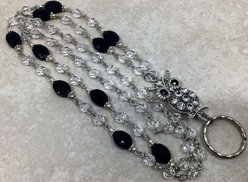 May include: Multi-strand beaded lanyard with black and clear beads.  The lanyard features an ornate owl-shaped badge holder with clear rhinestone accents. The metal chain and clasp are silver-toned.  The black beads are oval-shaped and matte. The clear beads are faceted and sparkly. This is a stylish and functional accessory for holding ID cards or keys.