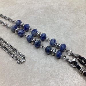 May include: Eyeglass chain featuring a silver-tone chain and blue sodalite beads. The chain has a double strand design with a series of blue beads and silver accents. The chain ends with black rubber loops for attaching to glasses.