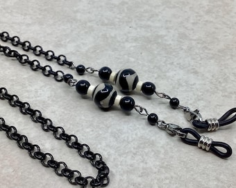 May include: Black eyeglass chain with small, round links. The chain has decorative beads with a black and white pattern, and small black beads. The chain ends with black rubber loops to hold the glasses.
