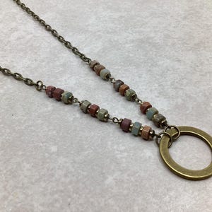 Puede incluir: Un collar con una cadena de color bronce y un gran colgante circular. La cadena está adornada con pequeñas cuentas de colores en tonos rojos, verdes y marrones. El colgante es un círculo abierto simple.